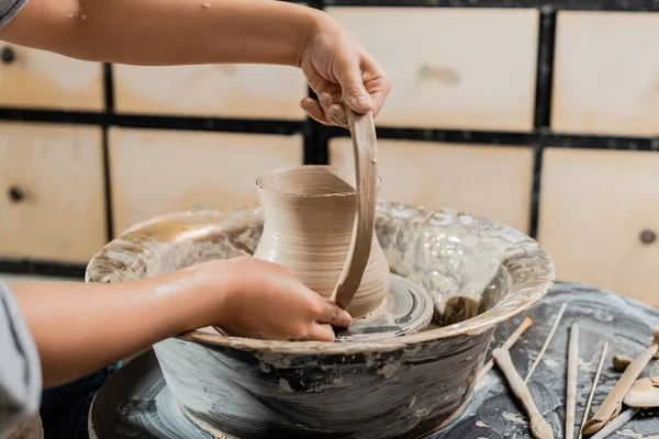 Обрізаний вид на молоду жінку-кераміка, що робить глиняний глечик і працює з гончарним колесом біля дерев'яних інструментів у розмитій художній майстерні, виробництво і процес вирощування кераміки — стокове фото