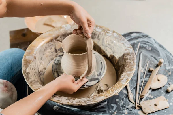 Vue en grand angle du jeune potier féminin en vêtements de travail créant une cruche en argile et travaillant avec une roue de poterie près d'outils en bois sur la table dans un atelier d'art, la production et le processus de poterie artisanale — Photo de stock