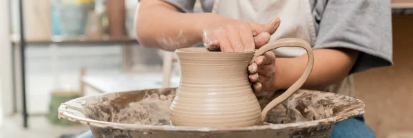 Cropped view of blurred young female potter in apron and workwear making clay jug while working with pottery wheel in art studio at background, clay shaping technique and process, banner — Stock Photo