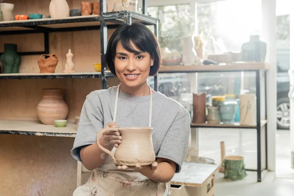 Fröhliche junge asiatische Kunsthandwerkerin in Schürze hält Tonkanne in der Hand und blickt in die Kamera, während sie in einer verschwommenen Keramikwerkstatt im Hintergrund arbeitet, Ton formt Technik und Prozess — Stockfoto
