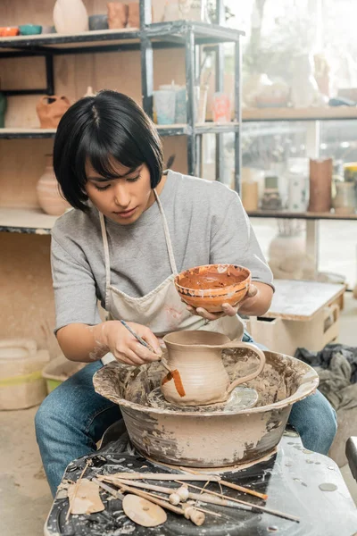 Молода брюнетка азіатська майстриня в фартухах малює на глиняному глечику на гончарному колесі, працюючи біля дерев'яних інструментів на столі в керамічній майстерні на фоні, технологія формування глини та процес — стокове фото
