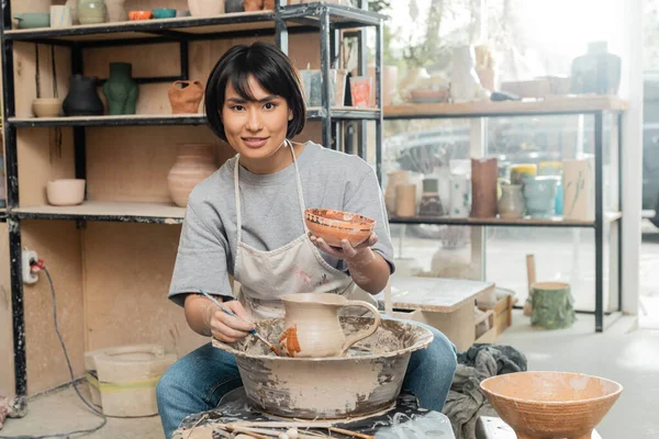 Joven artesana asiática alegre en delantal pintando jarra de arcilla y mirando a la cámara cerca de rueda de cerámica y herramientas de madera en taller de arte en el fondo, técnica y proceso de conformación de arcilla - foto de stock