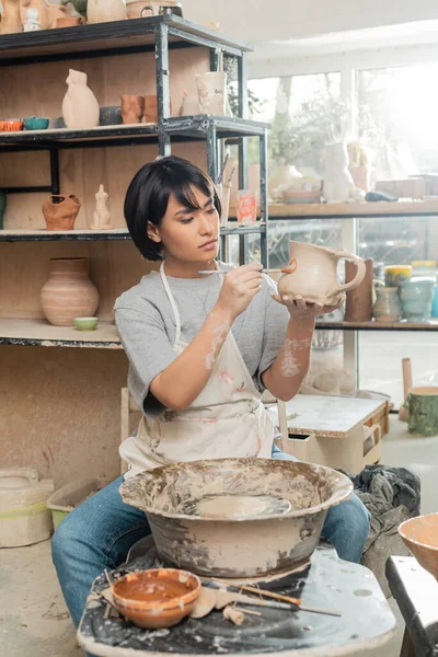 Young asian artisan in apron painting on clay jug while working near pottery wheel and wooden tools on table in ceramic workshop at blurred background, clay shaping technique and process — Stock Photo