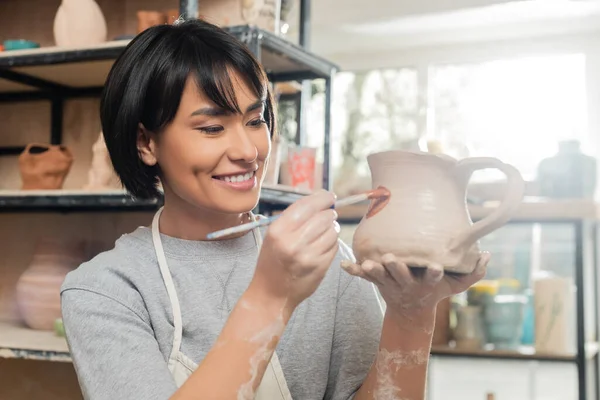 Portrait of cheerful young brunette asian artisan in apron and workwear painting on clay jug and working in blurred ceramic workshop at background, clay shaping technique and process — Stock Photo