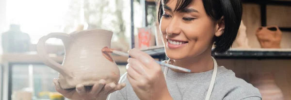 Lächelnde junge asiatische Kunsthandwerkerin, die auf Tonkannen malt, während sie im verschwommenen Keramik-Atelier im Hintergrund arbeitet, Tonformtechnik und -verfahren, Banner — Stockfoto
