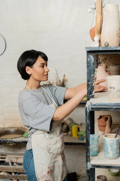Улыбающаяся молодая азиатская ремесленница в фартуке кладет керамическую скульптуру на полку на стойку, стоя в размытом художественном цехе на заднем плане, технику формирования глины и процесс — стоковое фото
