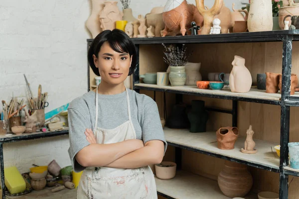 Lächelnde junge asiatische Kunsthandwerkerin in Schürze, die die Arme verschränkt und in die Kamera blickt, während sie in der Nähe von Regalen mit Keramikskulpturen in der Kunstwerkstatt steht, Töpferatelier-Szene — Stockfoto