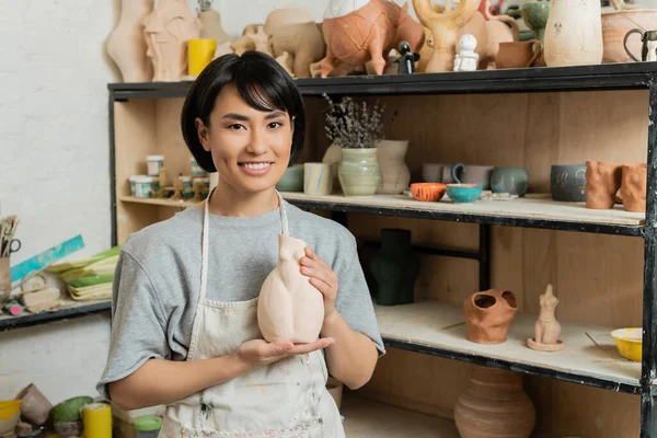 Retrato de alegre jovem artesão asiático em avental segurando escultura de cerâmica e olhando para a câmera perto de rack em oficina de arte borrada no fundo, cena de estúdio de cerâmica — Fotografia de Stock