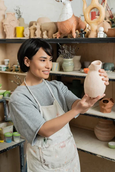 Sorridente giovane asiatica artigianale femminile in grembiule e abbigliamento da lavoro guardando scultura in ceramica mentre in piedi vicino a rack in laboratorio di ceramica offuscata sullo sfondo, scena studio di ceramica — Foto stock