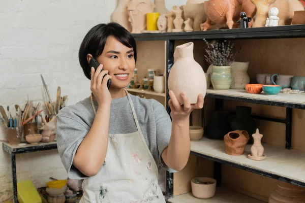 Lächelnde junge asiatische Kunsthandwerkerin in Schürze und Arbeitskleidung, die mit dem Smartphone spricht und im Hintergrund Keramikskulpturen in der Nähe von Regalen im verschwommenen Kunstatelier hält, Töpferatelier-Szene — Stockfoto