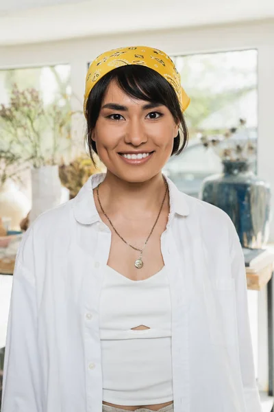 Porträt einer jungen lächelnden asiatischen Kunsthandwerkerin in Kopftuch und Arbeitskleidung, die in die Kamera blickt und im Hintergrund in einer unscharfen Keramikwerkstatt steht, Szene aus dem Töpferatelier — Stockfoto