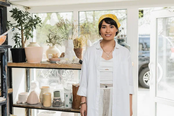 Portrait of smiling young asian female artisan in workwear and headscarf looking at camera while standing near ceramic sculptures at background in art workshop, pottery studio scene — Stock Photo