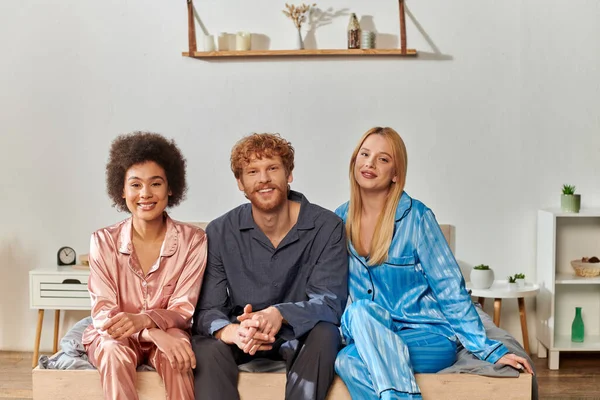 Open relationship, polygamy, understanding, three adults, redhead man and multicultural women in pajamas sitting on bed at home, cultural diversity, acceptance, bisexual — Stock Photo