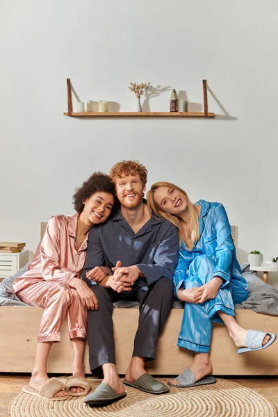 Open relationship concept, polygamy, understanding, three adults, happy man and multicultural women in pajamas sitting on bed at home, cultural diversity, acceptance, bisexual — Stock Photo