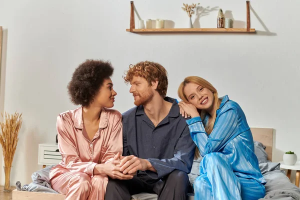 Полігамія концепція, розуміння, три дорослі, рудий чоловік і багатокультурні жінки в піжамах сидять на ліжку вдома, культурне різноманіття, прийняття, бісексуальні, позитивні, відкриті відносини — стокове фото