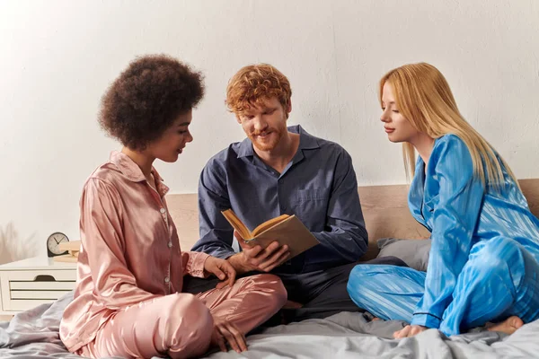 Concepto de relación abierta, hombre pelirroja lectura libro cerca de mujeres multiculturales felices en pijama sentado en la cama en casa, diversidad cultural, bisexual, poligamia, comprensión, tres adultos - foto de stock