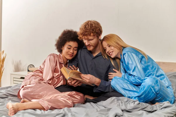 Концепция открытых отношений, рыжеволосый мужчина, читающий книгу рядом со счастливыми многокультурными женщинами в пижамах, сидящими дома на кровати, культурное разнообразие, бисексуализм, полигамия, взаимопонимание, трое взрослых — стоковое фото