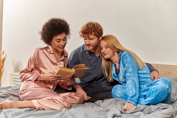 Концепция полигамии, африканская американка читает книгу рядом с любовниками в пижаме сидя на кровати дома, культурное разнообразие, бисексуалы, открытые отношения, понимание, три взрослых — стоковое фото