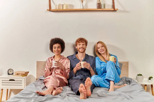 Concepto familiar no tradicional, poligamia, pelirroja y mujeres interracial en pijama sosteniendo tazas de café, rutina matutina, bisexual, comprensión, tres adultos, diversidad cultural, aceptación - foto de stock