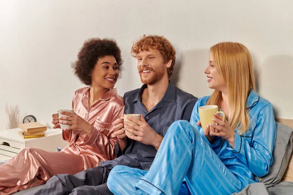 Libertad en concepto de relación, poligamia, hombre alegre y mujeres multiculturales en pijama sosteniendo tazas de café, rutina matutina, bisexual, comprensión, tres adultos, diversidad cultural - foto de stock