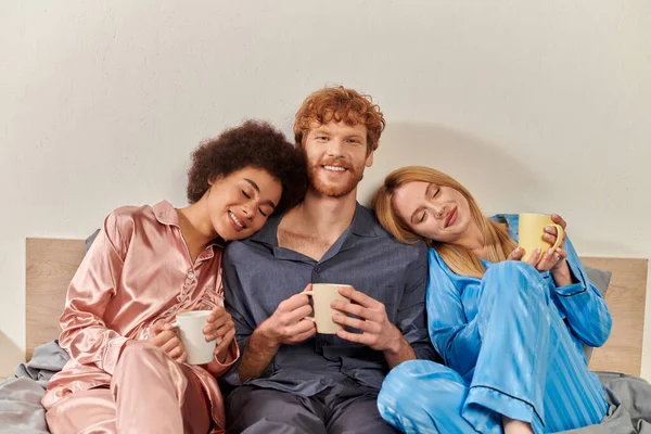 Открытая концепция отношений, полигамия, рыжий мужчина и мультикультурные женщины в пижаме с чашками кофе, утренняя рутина, бисексуальность, понимание, три взрослых, культурное разнообразие, принятие — стоковое фото