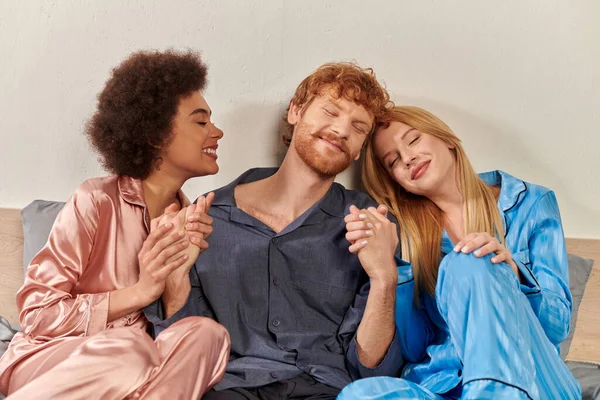 Open relationship, polygamy, happy three adults, redhead man and multicultural women in pajamas holding hands in bed, cultural diversity, acceptance, bisexual, modern family — Stock Photo