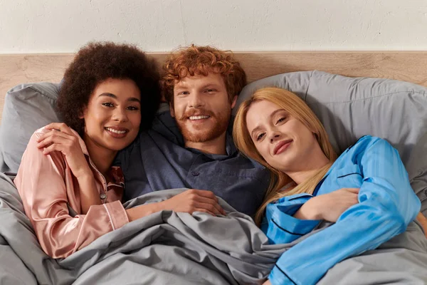 Polygamy, alternative relationships, three adults, happy redhead man and multicultural women lying under blanket together, cultural diversity, acceptance, bisexual, open relationship — Stock Photo