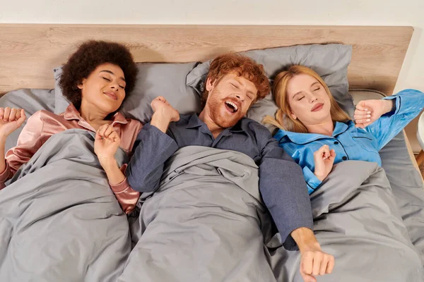 Polyamory concept, three adults, man and interracial women in pajamas waking up together, morning, under blanket, bedroom, cultural diversity, bisexual, open relationship, polygamy, top view — Stock Photo