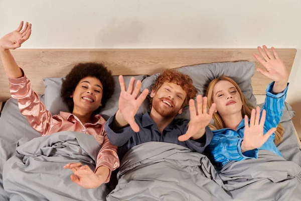 Polyamory concept, happy man and interracial women in pajamas waking up together, morning, under blanket, bedroom, diversity, bisexual, open relationship, polygamy, top view, outstretched hands — Stock Photo
