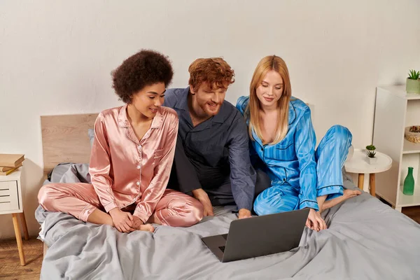 Relacionamento aberto, poligamia, compreensão, três adultos, ruivo homem e multiculturais mulheres em pijama assistindo filme no laptop, quarto, diversidade cultural, aceitação, bissexual — Fotografia de Stock