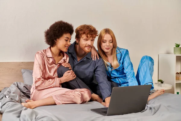 Poligamia, compreensão, três adultos, ruivo homem e multiculturais mulheres de pijama assistindo filme no laptop, quarto, diversidade cultural, aceitação, bissexual, relacionamento aberto — Fotografia de Stock