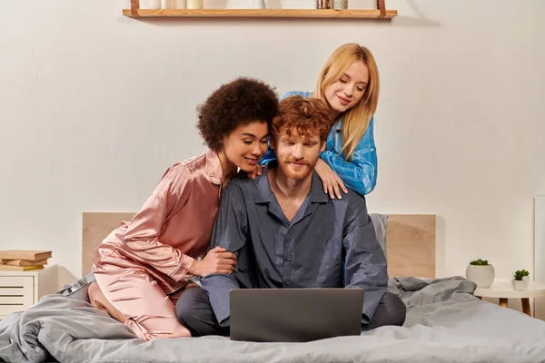 Принятие, открытые отношения, полигамия, понимание, три взрослых, рыжий мужчина и мультикультурные бисексуальные женщины в пижамах смотреть фильм на ноутбуке, спальня, культурное разнообразие — стоковое фото