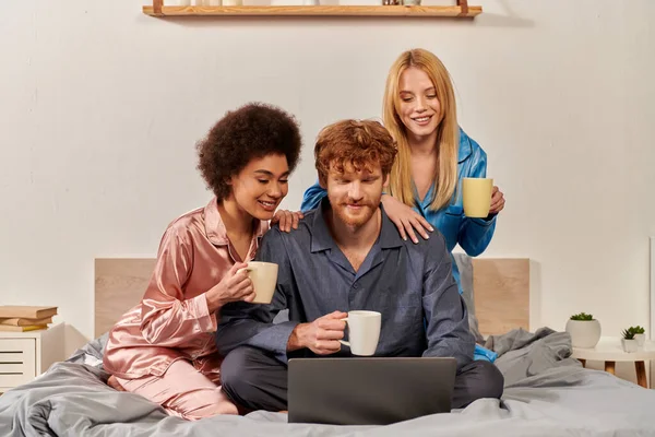 Polygamy, multicultural women and redhead man in pajamas watching movie on laptop, holding cups of coffee in bedroom, cultural diversity, acceptance, bisexual, open relationship — Stock Photo