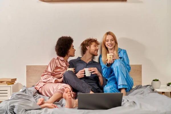 Polyamory relationships, multicultural women and redhead man in pajamas watching movie on laptop, holding cups of coffee in bedroom, cultural diversity, acceptance, bisexual, open relationship — Stock Photo
