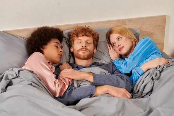 Полигамия, понимание, трое взрослых спят вместе, рыжий мужчина и мультикультурные женщины в пижамах, спальня, культурное разнообразие, принятие, бисексуальность — стоковое фото