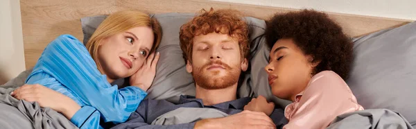 Любовний трикутник, полігамія, три дорослі сплять разом, рудий чоловік і мультикультурні жінки в піжамі, спальні, культурне різноманіття, прийняття, бісексуали, банер — стокове фото