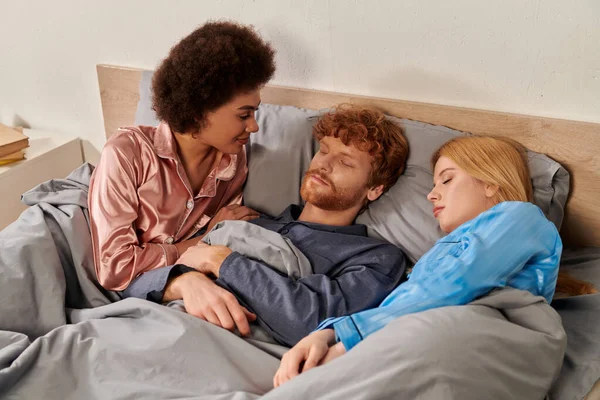 Любовный треугольник, проснувшаяся африканская американка смотрит на рыжего мужчину рядом с блондинкой партнер в постели, полиамори, нетрадиционные отношения, многорасовое, культурное разнообразие — стоковое фото