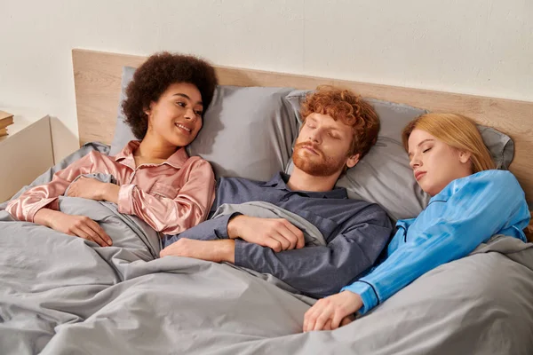 Love triangle, happy african american woman looking at sleepy redhead man near blonde female partner in bed, polyamory, non traditional relationships, multiracial, cultural diversity — Stock Photo