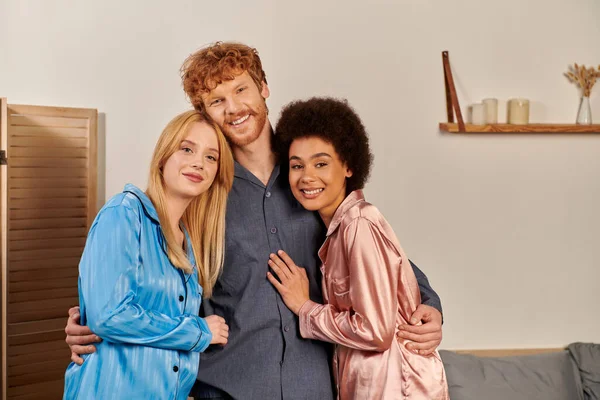 Polyamory lovers in pajamas, happy man with red hair hugging interracial women at home, cultural diversity, non traditional partners, freedom in relationship, acceptance and understanding, happiness — Stock Photo
