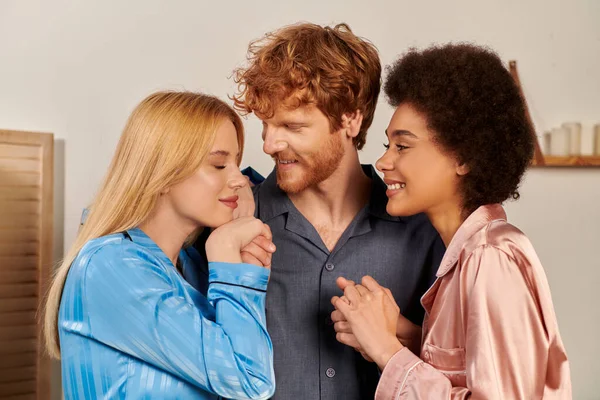 Polyamory lovers in pajamas, cheerful man with red hair hugging interracial women at home, cultural diversity, non traditional partners, freedom in relationship, acceptance and understanding — Stock Photo