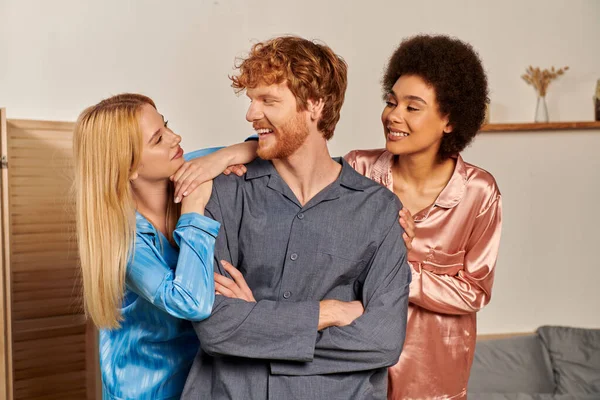 Polyamory lovers, happy man with folded arms near interracial women in pajamas, cultural diversity, non traditional partners, freedom in relationship, acceptance and understanding, joy — Stock Photo