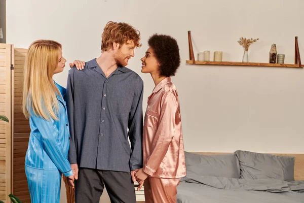 Polygamy lovers in pajamas, happy man with red hair holding hands of interracial women at home, non traditional partners, freedom in relationship, acceptance and understanding — Stock Photo