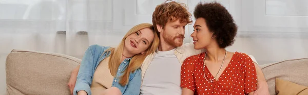 Modern family, polygamy concept, freedom in relationship, cultural diversity, redhead man sitting with multicultural women on couch in living room, polyamorous lifestyle, non traditional, banner — Stock Photo