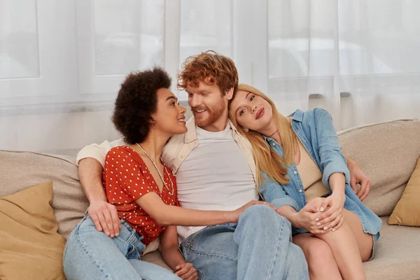 Polygamy concept, non traditional lovers, freedom in relationship, cultural diversity, redhead man sitting with multicultural women on couch in living room, polyamorous lifestyle, modern family — Stock Photo