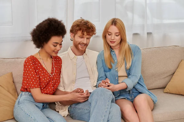 Modern family, polygamy concept, freedom in relationship, cultural diversity, redhead man holding hands with multicultural women, sitting on couch in living room, polyamorous lifestyle — Stock Photo