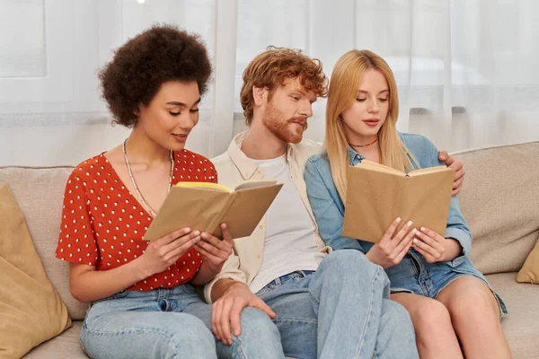 Conceito de poligamia, mulheres multiculturais inteligentes lendo livros perto de namorado na sala de estar, família moderna, triângulo amoroso, passatempo e lazer, liberdade no relacionamento — Fotografia de Stock