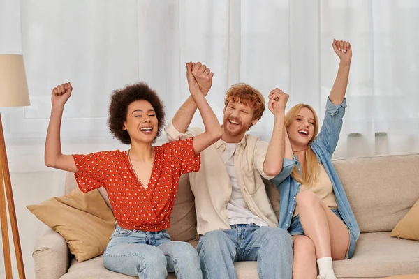 Polyamorous relationship, cultural diversity, redhead man sitting on couch with happy multiracial female lovers, raised hands, freedom and acceptance, love triangle, people in open relationship — Stock Photo