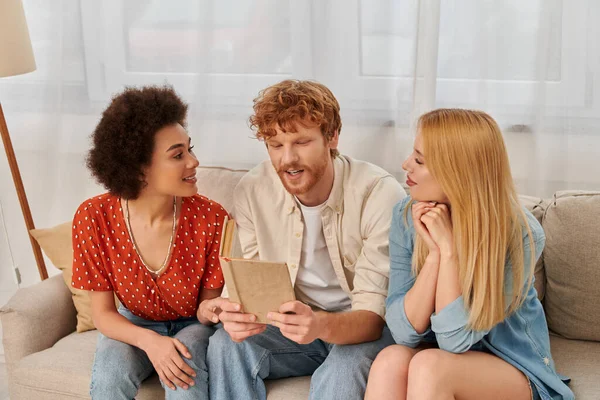 Open relationships, redhead man reading book to multicultural women in living room, non traditional lovers, cultural diversity, romance and love, modern family and polygamy concept — Stock Photo