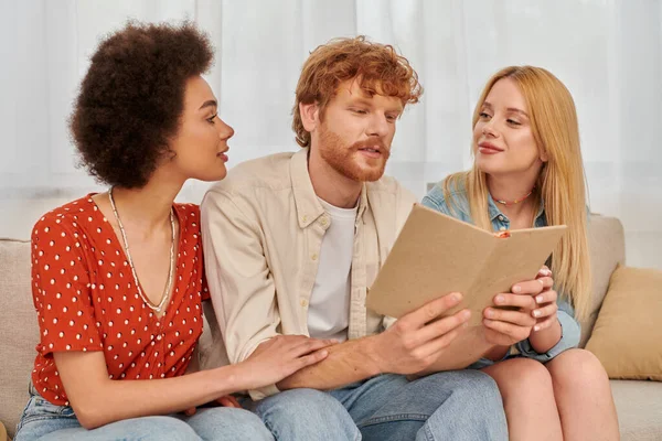 Open relationships, redhead man reading book to beautiful multicultural women in living room, non traditional lovers, cultural diversity, romance and love, modern family and polygamy concept — Stock Photo