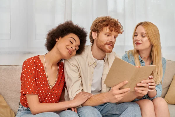 Polygamy, open relationships, redhead man reading book to pleased multicultural women in living room, non traditional lovers, cultural diversity, romance and love, modern family concept — Stock Photo